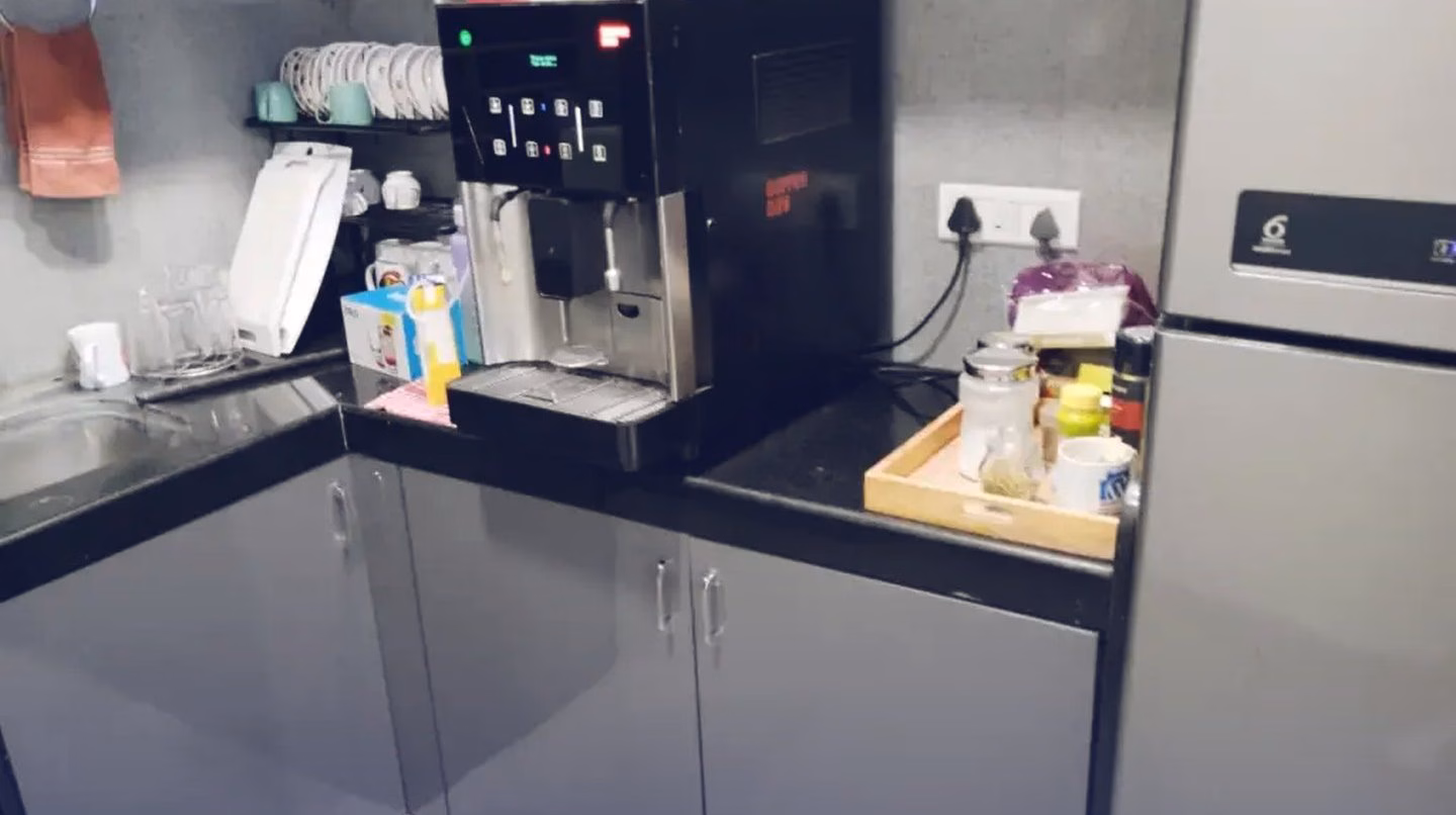The office pantry: a coffee machine on a black counter, cups on a rack, and a tray of snacks beside a tall fridge.