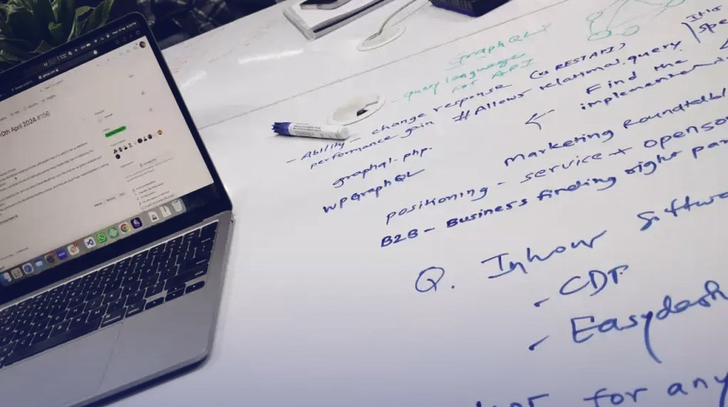 A laptop on a white desk beside handwritten notes on GraphQL and marketing, written straight onto the desk.