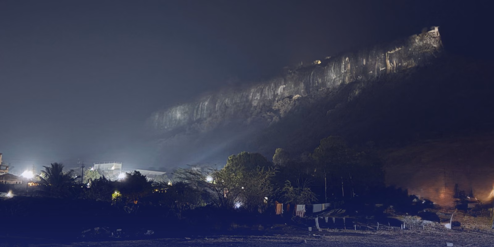 Shivneri fort at night, its cliff lit from below above the lights of Junnar.