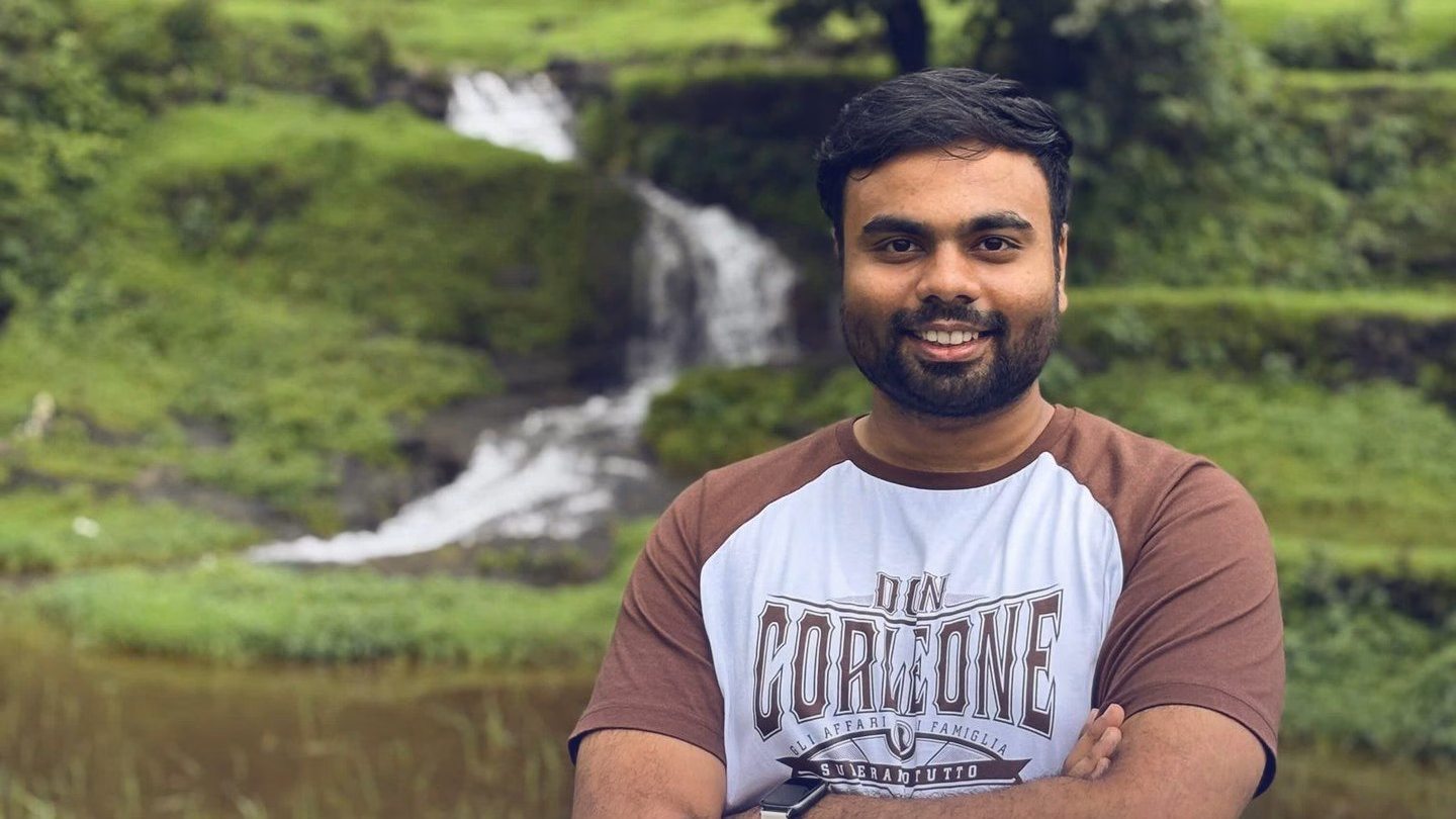 Abhinav smiling in front of a small waterfall on a green hillside.