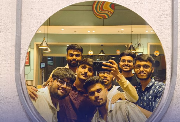 Seven friends crowd into a round mirror for a group selfie in a cafe.