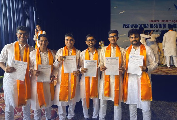 Six graduates in white kurtas and orange stoles hold their certificates on stage at Vishwakarma Institute.
