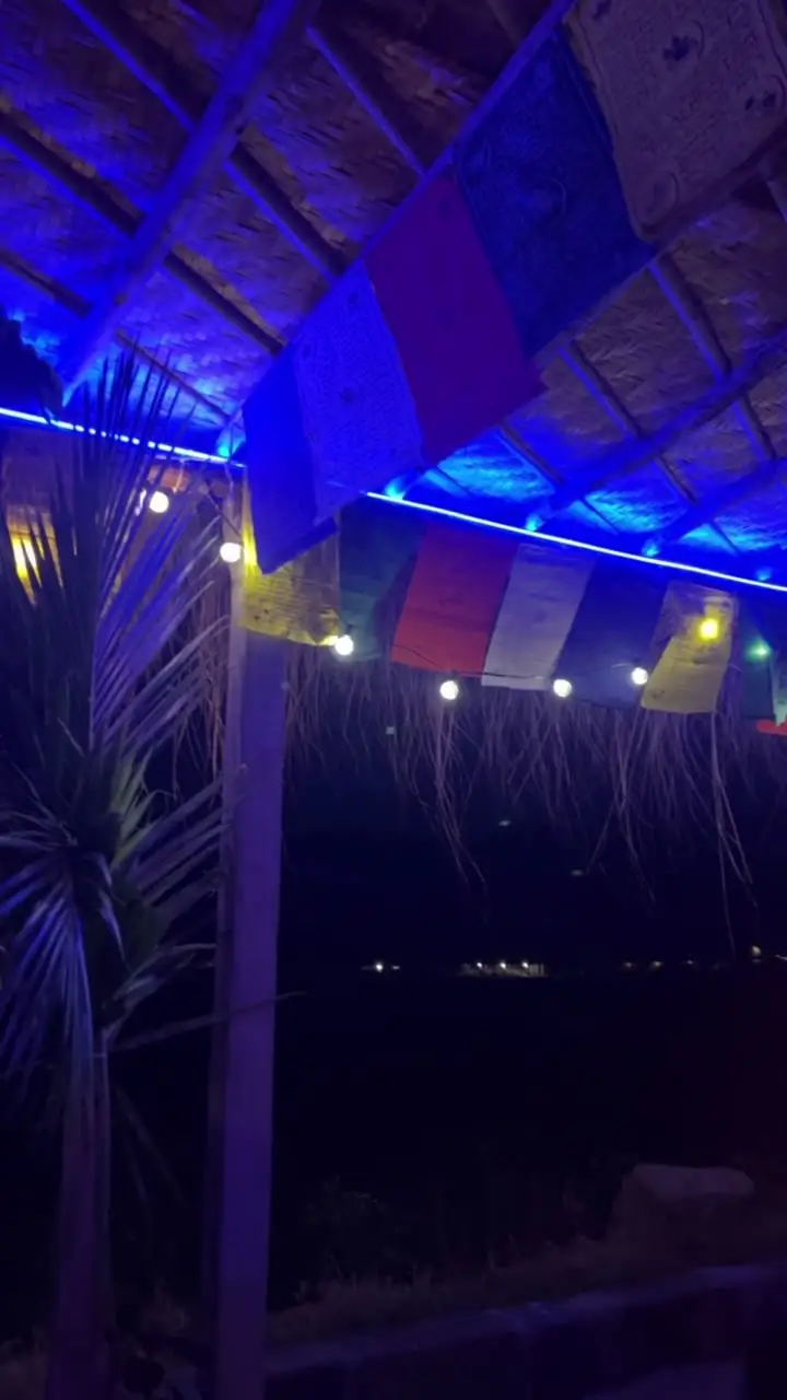 A café in Hampi at night: a bamboo roof lit blue, prayer flags, string lights and palm fronds against the dark. Chalk doodles draw in over it: a turmeric crescent moon, a few stars, a swag of fairy lights and the words Hampi after dark.